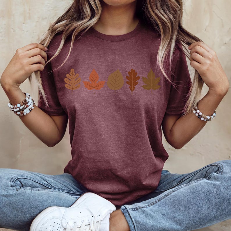 A woman wearing a maroon t-shirt with a graphic of autumn leaves, sitting cross-legged with her hands behind her head.