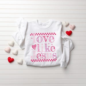 Puede incluir: Sudadera blanca con la frase "Love like Jesus" en letras rosas. El dise&ntilde;o incluye un s&iacute;mbolo de coraz&oacute;n y un borde a cuadros. Varias decoraciones en forma de coraz&oacute;n est&aacute;n dispersas alrededor de la sudadera.