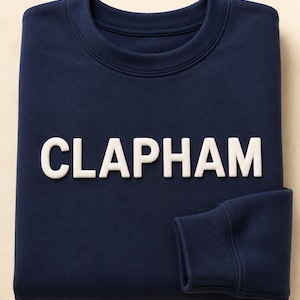 May include: A navy blue crewneck sweatshirt featuring the word CLAPHAM in raised, white, block-style lettering across the chest. The garment has long sleeves with ribbed cuffs and a ribbed neckline, constructed from a solid-colored fabric.