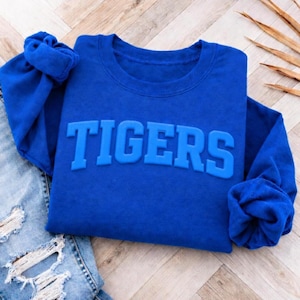 May include: A royal blue long-sleeve sweatshirt featuring the word TIGERS in raised, matching blue block lettering across the chest. The garment has a crew neckline and ribbed cuffs on the sleeves, providing a consistent monochromatic appearance.