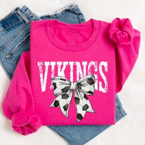 A pink sweatshirt with the word "VIKINGS" printed on it, along with a soccer ball bow design, is displayed on a bed.