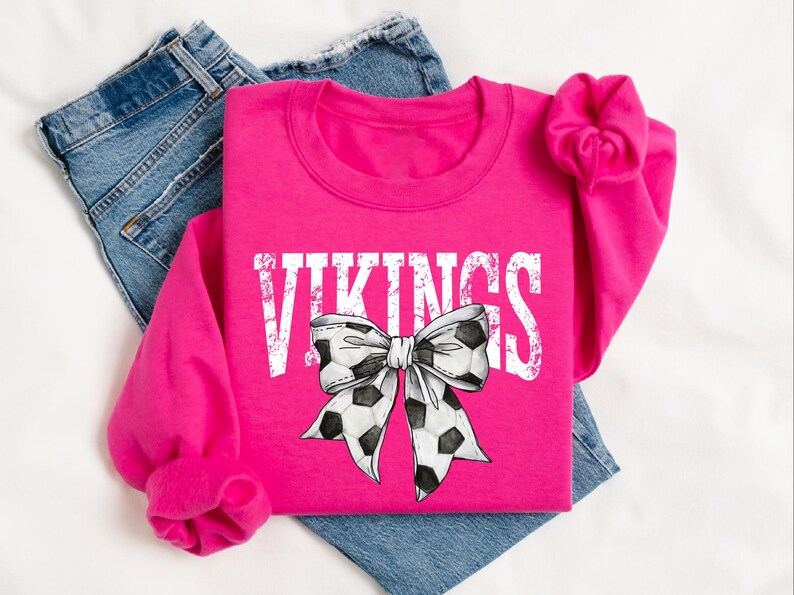 A pink sweatshirt with the word "VIKINGS" printed on it, along with a soccer ball bow design, is displayed on a bed.