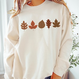 A woman wearing a white sweatshirt with a graphic design of autumn leaves on it.