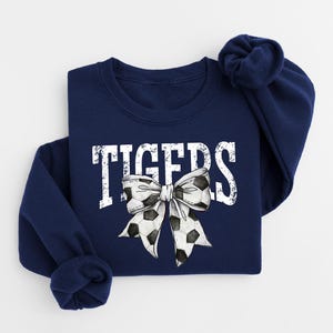 A navy blue sweatshirt with the word "TIGERS" printed on it, along with a large white bow featuring soccer balls.