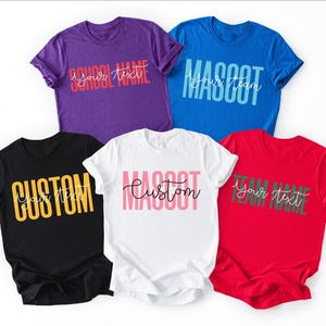 May include: Five short-sleeved crew neck t-shirts in purple, blue, black, white, and red. Each shirt features custom text printed in a combination of bold block lettering and a cursive script font, displaying phrases like SCHOOL NAME, MASCOT, and CUSTOM.