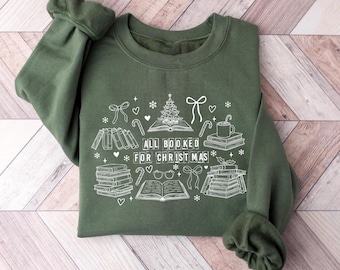 May include: An olive green sweatshirt with white graphic text and illustrations. The design includes stacks of books, a Christmas tree, candy canes, and the phrase "ALL BOOKED FOR CHRISTMAS".