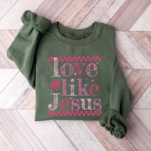 Puede incluir: Una sudadera verde oliva con el texto "Love like Jesus" en rosa y blanco. El texto est&aacute; estilizado con un coraz&oacute;n y detalles de tablero de ajedrez. La sudadera est&aacute; doblada sobre una superficie de madera clara marr&oacute;n y blanca.