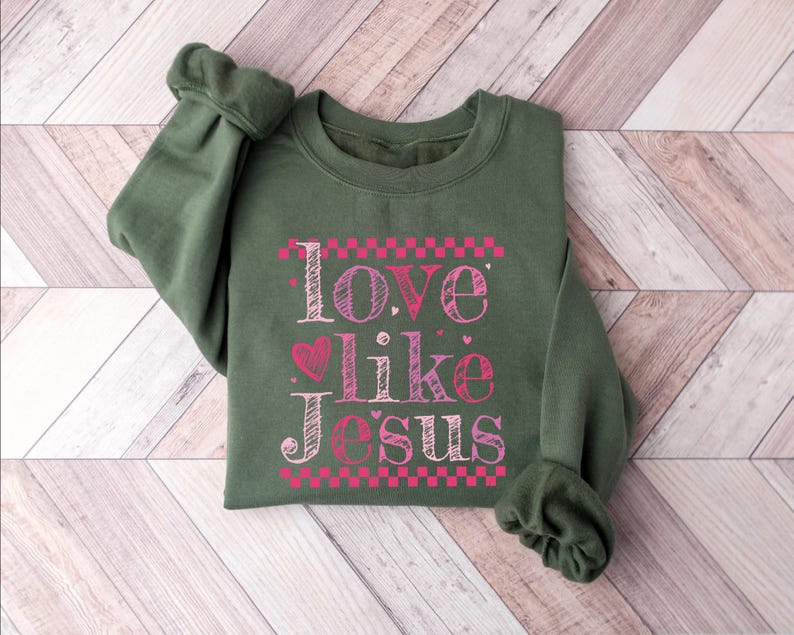 Puede incluir: Una sudadera verde oliva con el texto "Love like Jesus" en rosa y blanco. El texto est&aacute; estilizado con un coraz&oacute;n y detalles de tablero de ajedrez. La sudadera est&aacute; doblada sobre una superficie de madera clara marr&oacute;n y blanca.