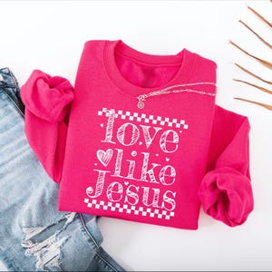 Puede incluir: Una sudadera rosa brillante con el texto blanco "Love like Jesus" y un coraz&oacute;n. Un collar plateado con un colgante descansa sobre la sudadera. Se combina con unos vaqueros azules.