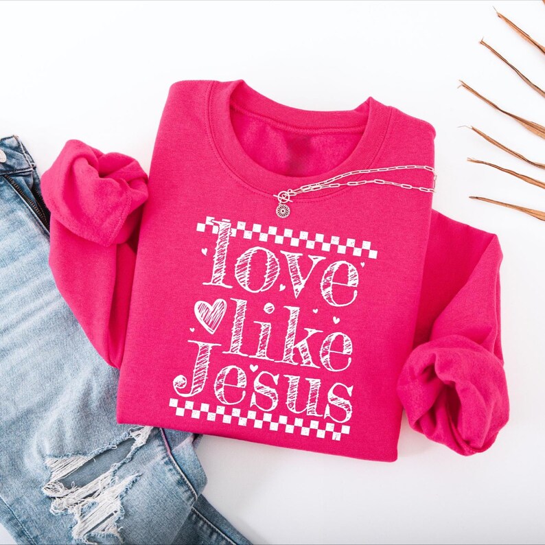 Puede incluir: Una sudadera rosa brillante con el texto blanco "Love like Jesus" y un coraz&oacute;n. Un collar plateado con un colgante descansa sobre la sudadera. Se combina con unos vaqueros azules.