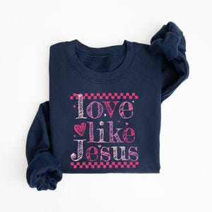 Puede incluir: Una sudadera azul marino con la frase "Love like Jesus" en escritura rosa. El texto incluye un coraz&oacute;n y est&aacute; enmarcado por patrones de cuadros rosas. La sudadera est&aacute; doblada sobre una superficie blanca.