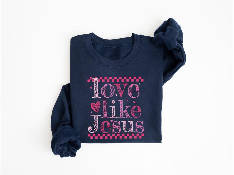 Puede incluir: Una sudadera azul marino con la frase "Love like Jesus" en escritura rosa. El texto incluye un coraz&oacute;n y est&aacute; enmarcado por patrones de cuadros rosas. La sudadera est&aacute; doblada sobre una superficie blanca.