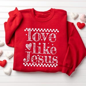 Puede incluir: Una sudadera roja con las palabras "Love like Jesus" en escritura blanca. El texto est&aacute; enmarcado por un patr&oacute;n de cuadros. Peque&ntilde;os corazones de fieltro en blanco, rosa y rojo est&aacute;n esparcidos alrededor.