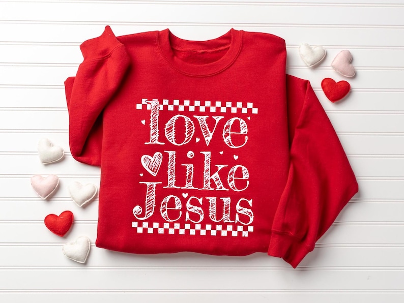 Puede incluir: Una sudadera roja con las palabras "Love like Jesus" en escritura blanca. El texto est&aacute; enmarcado por un patr&oacute;n de cuadros. Peque&ntilde;os corazones de fieltro en blanco, rosa y rojo est&aacute;n esparcidos alrededor.