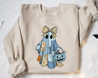 May include: Beige sweatshirt featuring a patchwork ghost design with a bow and a pumpkin basket. The ghost is made of blue, orange, and beige patches. The pumpkin is light blue with a carved face. Halloween-themed apparel.