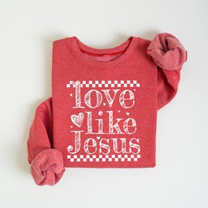 Puede incluir: Sudadera roja con la frase "Love like Jesus" en letras blancas. El texto est&aacute; centrado en la parte delantera de la prenda, con un coraz&oacute;n en lugar de la palabra "love". La sudadera tiene mangas largas y cuello redondo.