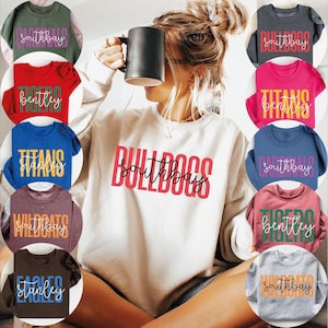 May include: A selection of crewneck sweatshirts and t-shirts in different colours, each displaying a team name and location. The central sweatshirt is white with red text reading "Bulldogs Southbay". Other shirts include "Tigers Bentley" and "Wildcats Southbay".
