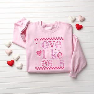 Puede incluir: Una sudadera rosa claro con la frase "Love like o's s" en rosa. El dise&ntilde;o incluye un coraz&oacute;n y patrones de tablero de ajedrez. Varios adornos en forma de coraz&oacute;n en rojo, rosa y blanco est&aacute;n dispersos alrededor de la sudadera.