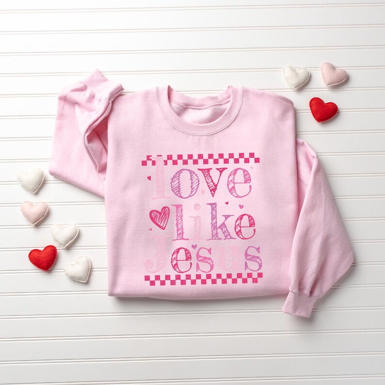 Puede incluir: Una sudadera rosa claro con la frase "Love like o's s" en rosa. El dise&ntilde;o incluye un coraz&oacute;n y patrones de tablero de ajedrez. Varios adornos en forma de coraz&oacute;n en rojo, rosa y blanco est&aacute;n dispersos alrededor de la sudadera.