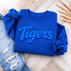 May include: A royal blue sweatshirt with the word "Tigers" in a raised, lighter blue script. The sweatshirt is displayed with a pair of distressed blue jeans on a light wood-grain surface. A dried palm leaf is in the background.