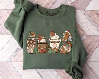 May include: Green sweatshirt featuring a festive design of gingerbread men and houses in various mugs and jars. The graphic includes gingerbread men, Christmas trees, and decorative elements, perfect for the holiday season.