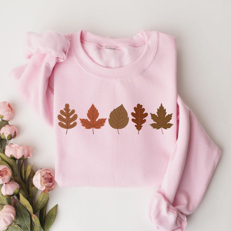 A pink sweatshirt with a row of autumn leaves printed on it, along with a bouquet of pink flowers to the side.