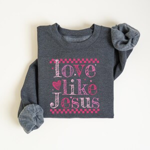 Puede incluir: Sudadera gris oscuro con la frase "Love like Jesus" en letras rosas y blancas. El texto est&aacute; enmarcado con un patr&oacute;n de cuadros rosas e incluye un coraz&oacute;n rosa. Las mangas est&aacute;n enrolladas.