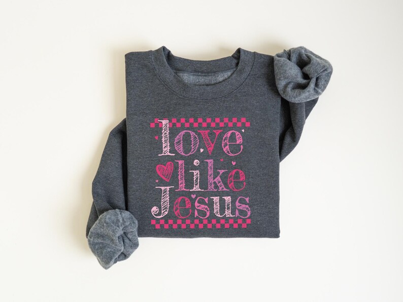 Puede incluir: Sudadera gris oscuro con la frase "Love like Jesus" en letras rosas y blancas. El texto est&aacute; enmarcado con un patr&oacute;n de cuadros rosas e incluye un coraz&oacute;n rosa. Las mangas est&aacute;n enrolladas.