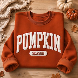 May include: A burnt orange crewneck sweatshirt featuring the white text PUMPKIN in a large arched varsity font, with the word SEASON printed in a smaller white rectangular block below. The garment has long sleeves and a ribbed collar.