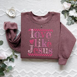 Puede incluir: Una sudadera granate con la frase "Love Like Jesus" en letras rosas y blancas. El dise&ntilde;o incluye corazones y un borde a cuadros. Se ve un peque&ntilde;o collar plateado en el escote.