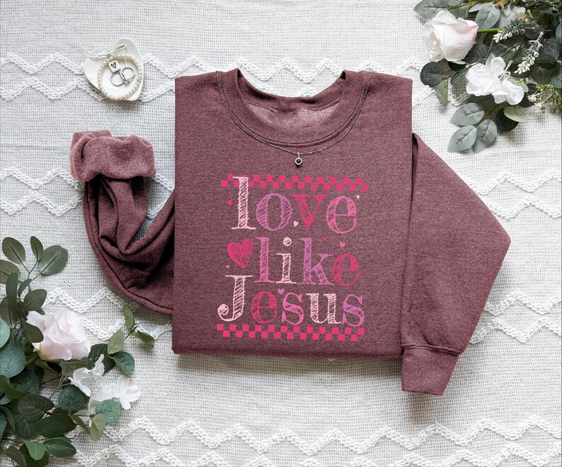 Puede incluir: Una sudadera granate con la frase "Love Like Jesus" en letras rosas y blancas. El dise&ntilde;o incluye corazones y un borde a cuadros. Se ve un peque&ntilde;o collar plateado en el escote.