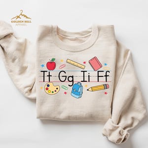 May include: A beige sweatshirt featuring a colourful graphic of school supplies and the letters "Tt Gg Ii Ff" on a lined paper background. The graphic includes a red apple, a ruler, a notebook, a paint palette, a backpack, and a pencil.