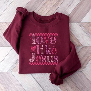 Puede incluir: Una sudadera burdeos con la frase "Love like Jesus" en un dise&ntilde;o rosa y blanco. El texto incluye un s&iacute;mbolo de coraz&oacute;n y est&aacute; enmarcado por un patr&oacute;n de cuadros rosas. La sudadera est&aacute; doblada sobre una superficie de madera clara.