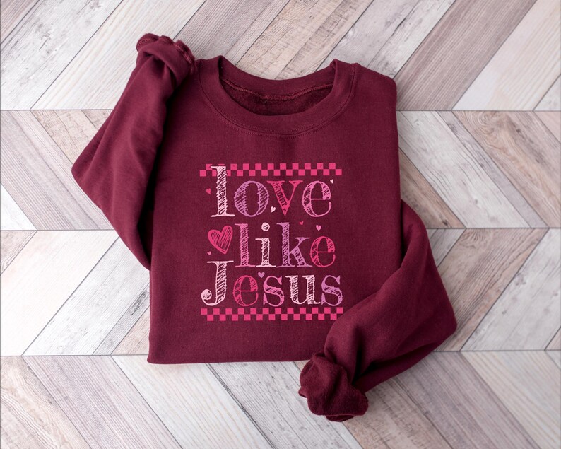 Puede incluir: Una sudadera burdeos con la frase "Love like Jesus" en un dise&ntilde;o rosa y blanco. El texto incluye un s&iacute;mbolo de coraz&oacute;n y est&aacute; enmarcado por un patr&oacute;n de cuadros rosas. La sudadera est&aacute; doblada sobre una superficie de madera clara.