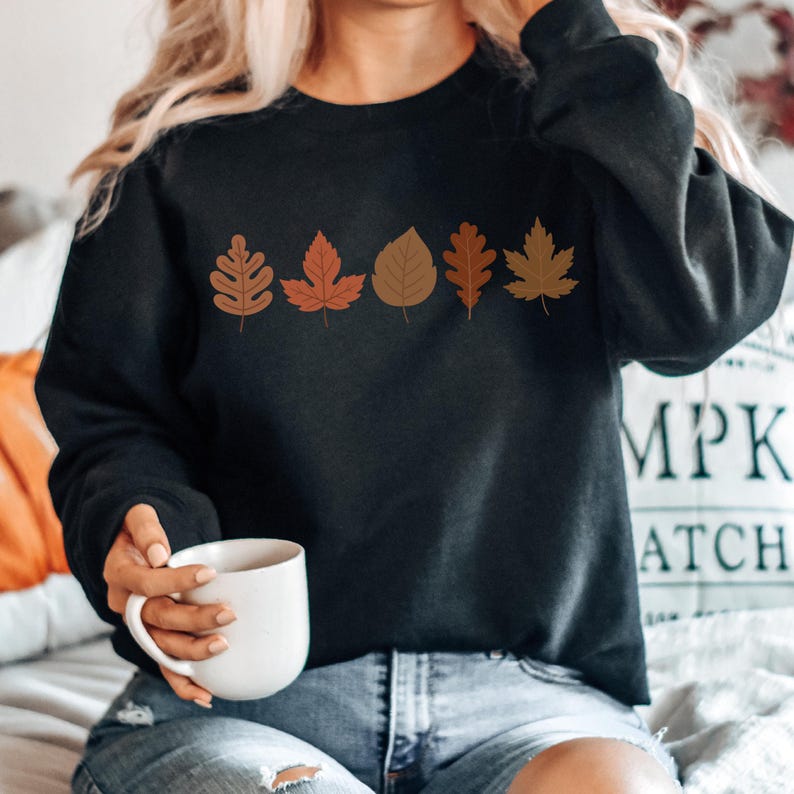 A woman wearing a black sweatshirt with a graphic of autumn leaves is holding a white mug and sitting on a bed.
