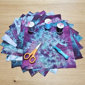 May include: A stack of square fabric pieces featuring a tie-dye pattern in shades of purple and teal. Included are three spools of thread in gray, light blue, and black, a pair of orange-handled scissors, and a light blue handled seam ripper.