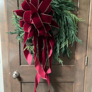 May include: A circular wreath made of dark green evergreen branches adorned with a large, multi-looped deep red velvet bow. The bow features long, flowing ribbon tails that hang downward, and the edges of the velvet are trimmed in gold.