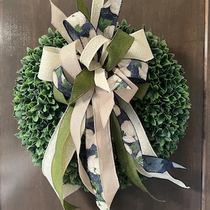 May include: A round green boxwood wreath adorned with a large decorative bow. The bow features ribbons in solid beige, textured olive green, and a navy blue fabric patterned with white pumpkins and green vines, creating a layered appearance.