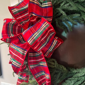May include: A festive Christmas wreath with a large red plaid bow. The bow features red, green, gold, and blue stripes. The wreath is made of green pine branches, creating a classic holiday decoration.
