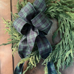 May include: A circular wreath constructed from dense green evergreen foliage, featuring a large, multi-looped decorative bow made of fabric with a dark green and navy blue plaid pattern, with two long ribbon tails extending downward.