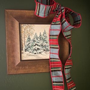 May include: A square framed painting of snow-covered pine trees in a winter forest, featuring a textured brown wooden frame. A large decorative fabric bow with a red, green, blue, and yellow plaid pattern is attached to the top corner.