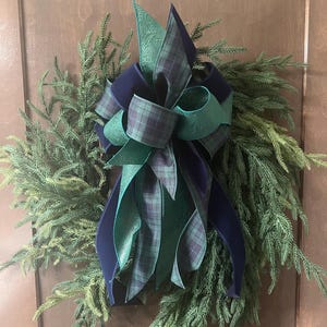 May include: A decorative wreath made of green pine needles featuring a large, multi-layered bow. The bow is constructed from ribbons in solid navy blue, a green and navy blue plaid pattern, and a solid teal green with an embossed floral texture.
