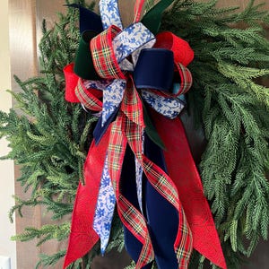 May include: A decorative wreath featuring a large multi-layered bow made of various ribbons. The ribbons include red fabric with a subtle embossed pattern, navy blue velvet, a red and green plaid, and a white ribbon with a blue floral print.