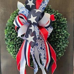 Op de afbeelding: Een decoratieve krans met een dichte groene basis, versierd met een patriottische strik. De strik bevat marineblauwe linten met witte sterren, rood-wit gestreepte linten en linten met een blauw bloemenpatroon. Geschikt voor deur- of wanddecoratie.