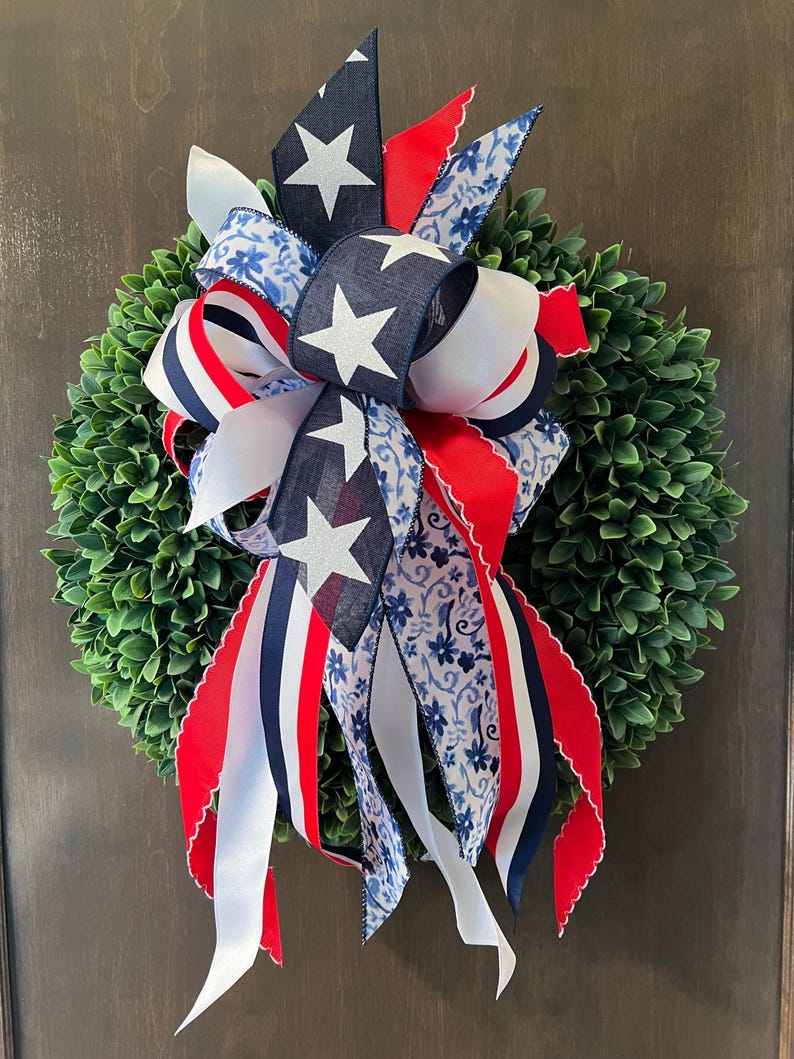Op de afbeelding: Een decoratieve krans met een dichte groene basis, versierd met een patriottische strik. De strik bevat marineblauwe linten met witte sterren, rood-wit gestreepte linten en linten met een blauw bloemenpatroon. Geschikt voor deur- of wanddecoratie.