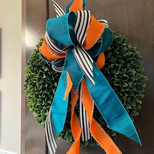 May include: A green boxwood wreath with a large bow made of teal, black and white striped, and orange ribbon. The bow has frayed edges.