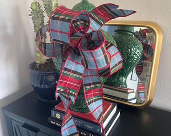 May include: A decorative bow made of fabric featuring a plaid pattern in shades of light blue, red, green, and yellow with a thin red wire-edged border. The bow has multiple loops and long, flowing tails that drape downward in a symmetrical arrangement.