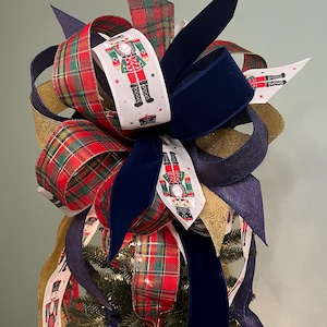 May include: A Christmas tree topper featuring a large bow with red and green plaid ribbon, navy blue velvet ribbon, and gold embossed ribbon. The bow is decorated with ribbons printed with a nutcracker soldier design.