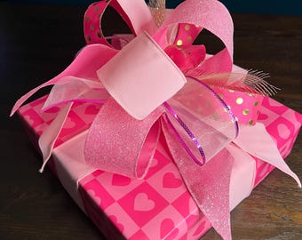 May include: A square gift box wrapped in pink paper with a heart pattern. The box is topped with a large, multi-layered bow featuring pink ribbons, some with gold polka dots and glitter accents. A decorative gift for any occasion.