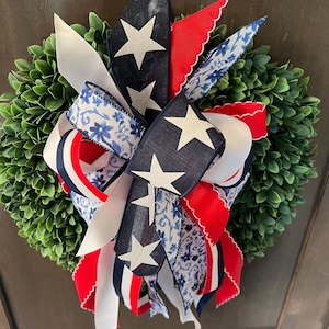 Op de afbeelding: Een decoratieve krans met een dichte groene basis, versierd met patriottische linten. De linten tonen wit, marineblauw met witte sterren, rood en blauwe bloempatronen. Geschikt voor deur- of wanddecoratie.